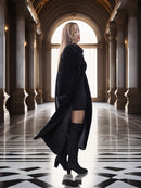 Woman in a long black coat standing in an elegant hallway with columns and large windows.