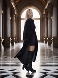 Woman in a long black coat standing in an elegant hallway with columns and large windows.