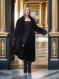 Woman in a black coat standing in an elegant interior setting with gold accents.