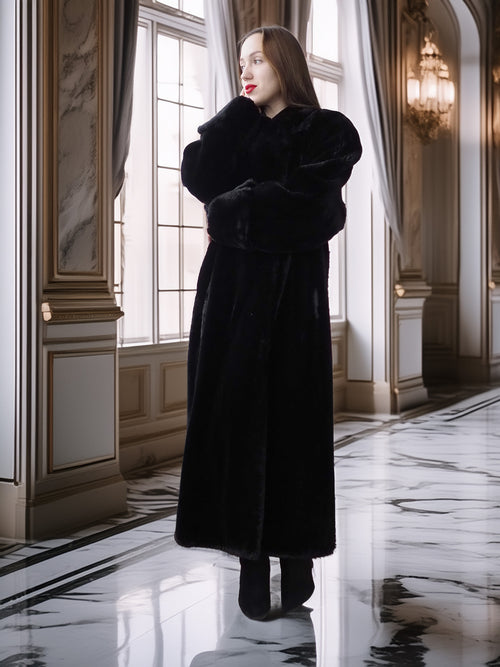Woman in a long black coat standing in an elegant interior setting with marble floors and large windows.