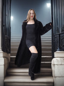 Woman in a long black coat and knee-high boots standing on steps at night.