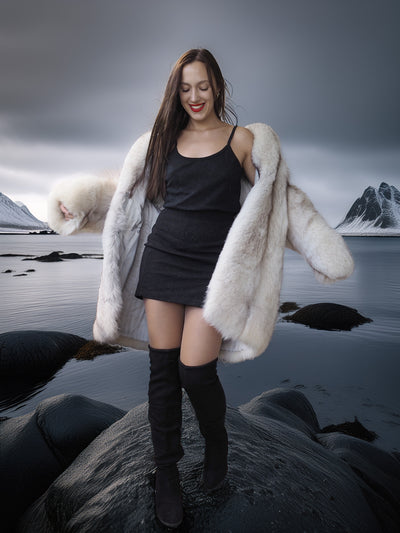 Woman in a white fur coat and black outfit standing on rocks with a mountainous landscape in the background