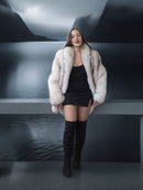 Woman in a fur coat and black dress standing by a lake with mountains in the background