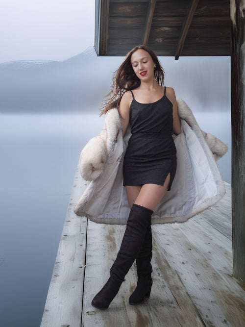 Woman in a black dress and fur coat standing on a wooden deck by a body of water.