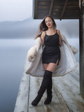Woman in a black dress and fur coat standing on a wooden deck by a body of water.