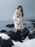 Woman in a fur coat standing on a snowy landscape with rocks and water.