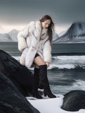 Woman in a white fur coat and black boots sitting on a rock by a snowy landscape with mountains.