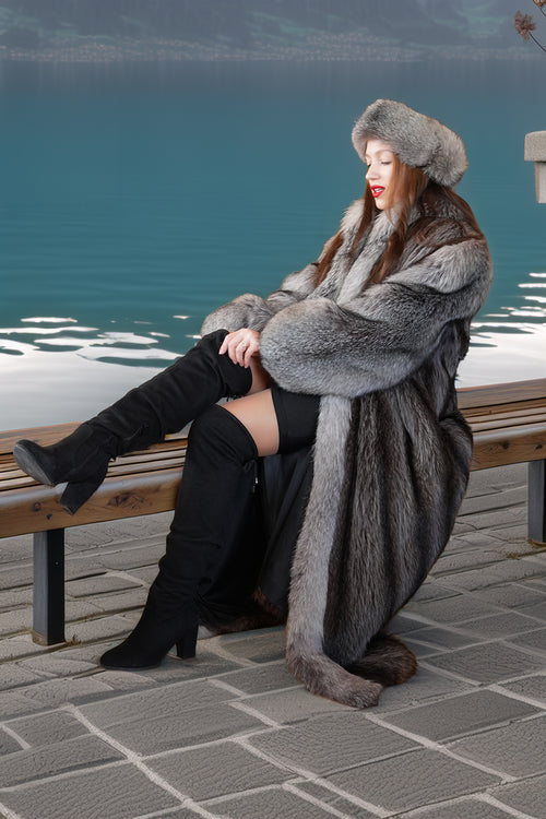 Woman in a fur coat and boots sitting on a bench by a body of water.