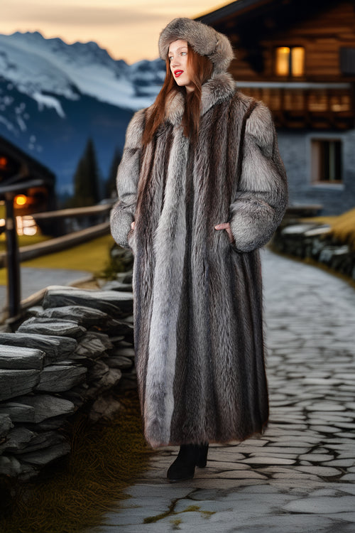 Woman in a fur coat standing in front of a mountain cabin at dusk.
