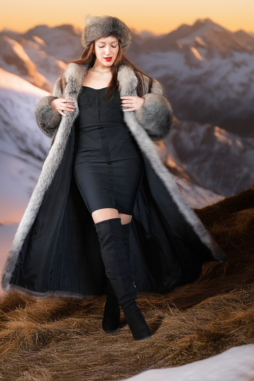Woman in a long fur coat and black outfit standing in a mountainous landscape.