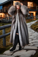 Person wearing a long fur coat and hat in front of a wooden cabin.