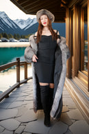 Woman in a long fur coat and black dress standing on a stone deck with mountains in the background
