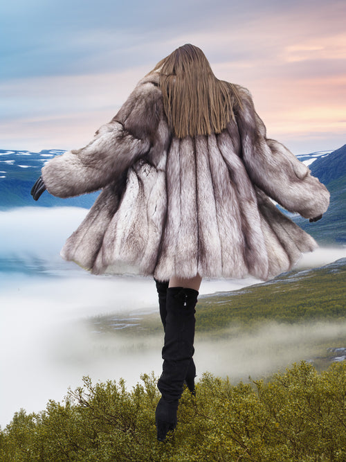 Back view of plush blue fox coat with fully let out pelts and wide silhouette, worn above a cloud-filled valley. Luxury fur outerwear.