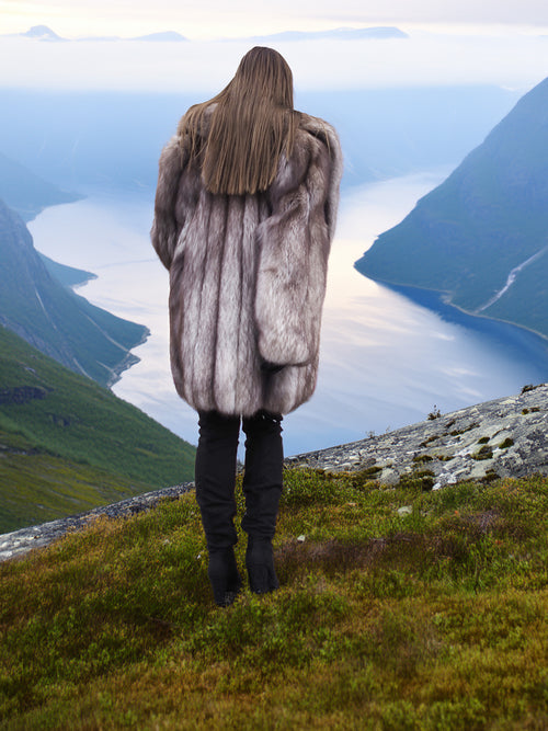 Back view of plush silver-blue fox fur coat with thick texture, worn above a fjord. Premium real fur jacket for winter elegance.
