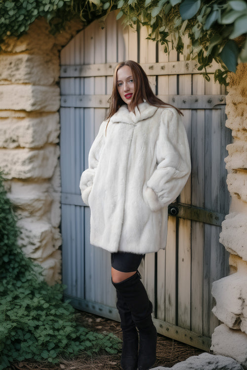 A woman wearing a voluminous white faux fur coat, black leggings, and black over-the-knee boots stands in front of a weathered gray wooden door framed by stone walls and green ivy.