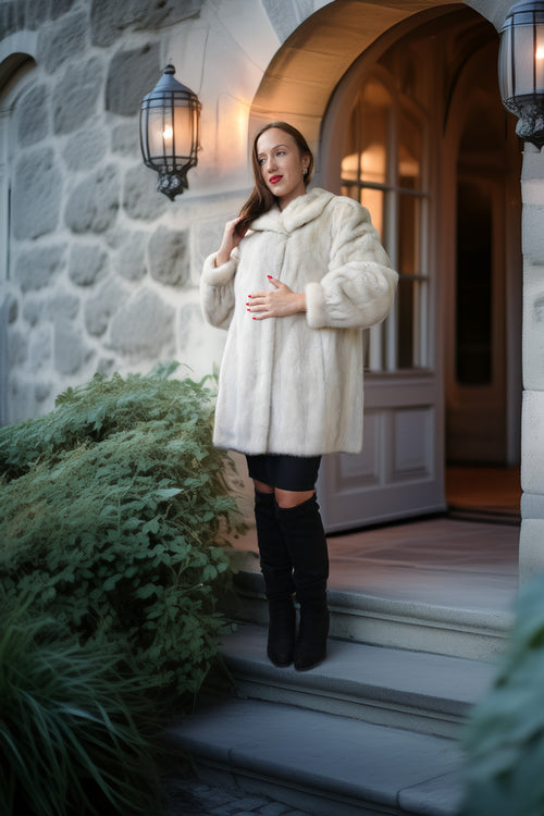 A woman stands on the steps of a stone building wearing a voluminous cream faux-fur coat, a black mini skirt, and black knee-high boots.