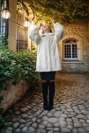 A woman wearing a voluminous white faux fur coat, black skirt, and black over-the-knee boots stands on a cobblestone path in front of a stone building with warm lights and greenery.