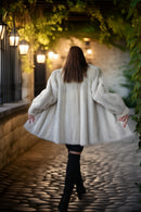 A woman viewed from behind wearing a voluminous white faux fur coat, black pants, and black boots, walking on a cobblestone path under a stone archway with hanging lanterns and green ivy.