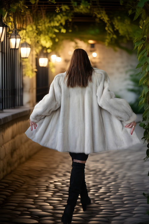 A woman viewed from behind wearing a voluminous white faux fur coat, black pants, and black boots, walking on a cobblestone path under a stone archway with hanging lanterns and green ivy.