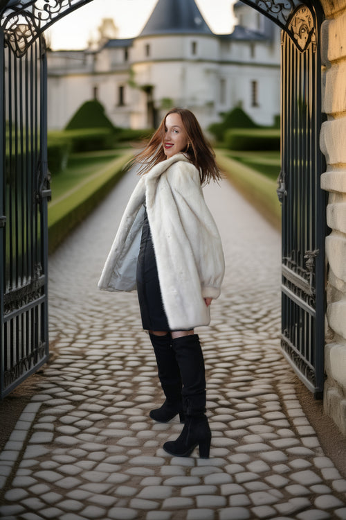 A woman stands in a stone courtyard framed by open black iron gates, wearing a voluminous off-white faux fur coat over a dark dress and black over-the-knee boots. A chateau and manicured hedges are visible in the background.
