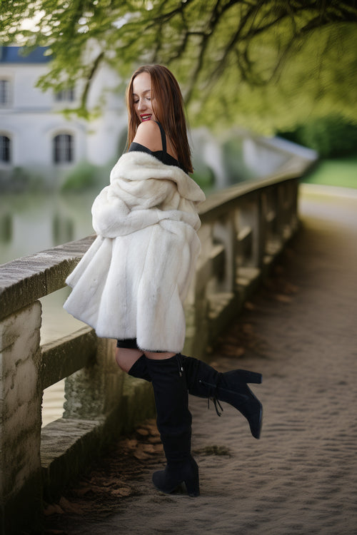 A woman posing on a stone bridge outdoors, wearing a voluminous white faux fur coat over a black outfit with black over-the-knee boots.