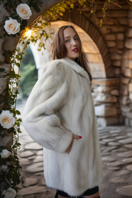 A woman wearing a voluminous white fur coat stands in a stone archway decorated with white roses and green vines.
