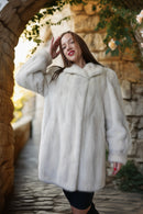 A woman wearing a plush white fur coat stands in a stone walkway under an archway with greenery.