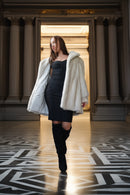 A woman walks through a grand hall with classical columns, wearing a black strapless dress, black thigh-high boots, and an oversized white faux-fur coat.