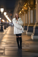 A woman wearing a white faux fur coat, black mini skirt, and black over-the-knee boots walks down a city street at night with warm streetlights and building columns in the background.