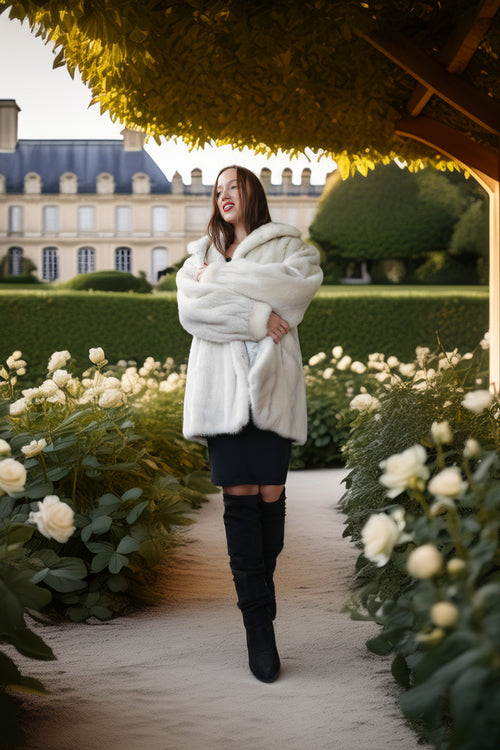 A woman stands on a garden path wearing a voluminous white faux fur coat, a black mini skirt, and black over-the-knee boots. The background features a grand classical building, manicured hedges, and white roses under a leafy canopy.