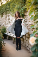 A woman stands outdoors on a stone path beside a stone wall with greenery, wearing a fitted black dress, a light gray coat, and black knee-high boots.