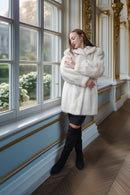 A woman stands by a large window in an ornate room, wearing a voluminous white faux fur coat, black leggings, and black over-the-knee boots.