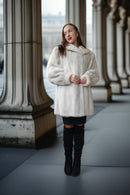 A woman wearing a fluffy white faux fur coat, black mini skirt, and black over-the-knee boots stands in a colonnade with large stone columns.