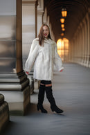 A woman wearing a voluminous white fur coat, black thigh-high cutout boots, and black knee-high socks walks through a covered outdoor corridor with warm hanging lights in the background.