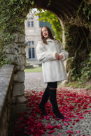 A woman wearing a white faux fur coat, black leggings, and black over-the-knee boots stands under a stone archway with red rose petals on the ground.