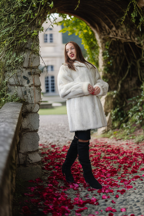 A woman wearing a white faux fur coat, black leggings, and black over-the-knee boots stands under a stone archway with red rose petals on the ground.