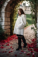 A woman wearing a voluminous white faux fur coat, black mini skirt, and black over-the-knee boots stands in a stone archway with red rose petals on a cobblestone path, with a castle in the background.