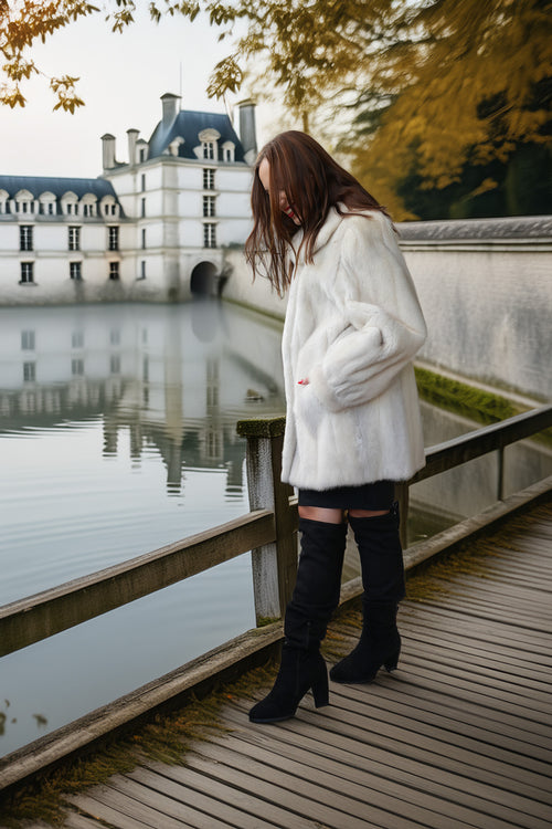 A woman stands on a wooden boardwalk beside a calm lake, wearing a voluminous white faux-fur coat, black mini skirt, and black over-the-knee boots. A large historic chateau is reflected in the water in the background.