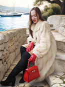 Woman in a white fur coat and red gloves holding a red handbag by a waterfront.