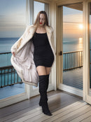 Woman in a black dress and white fur coat standing on a wooden deck with ocean view