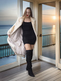Woman in a black dress and white fur coat standing on a wooden deck with ocean view