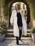 Woman in a long white fur coat standing in an archway with plants on either side.