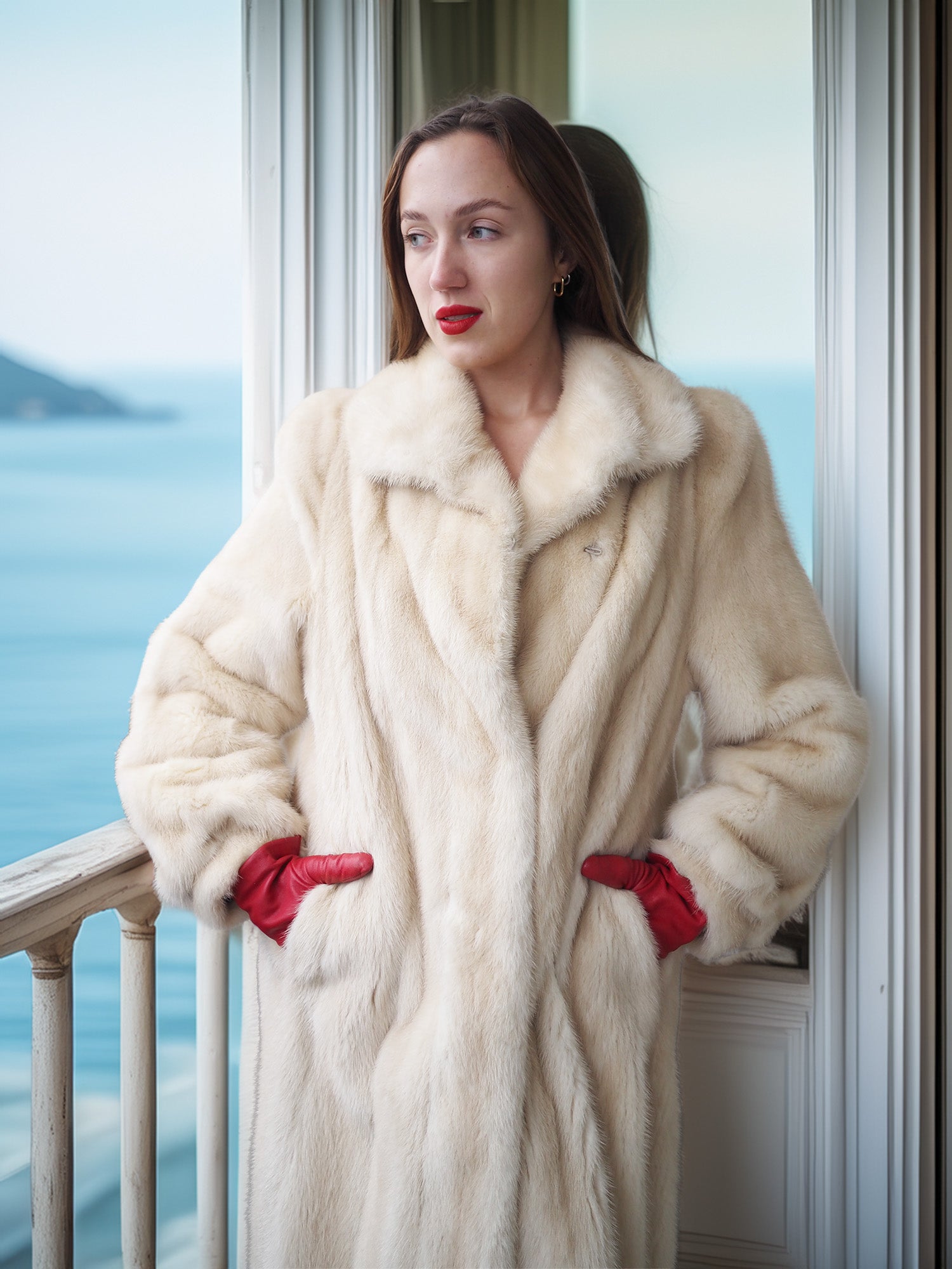 Woman wearing a white fur coat and red gloves standing on a balcony with a scenic view.