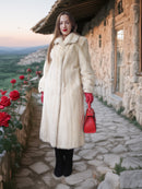 Woman in a fur coat holding a red handbag in a scenic outdoor setting with stone building and roses.