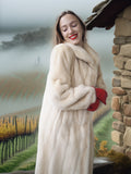 Woman in a fur coat and red gloves standing in front of a scenic background with a stone wall.