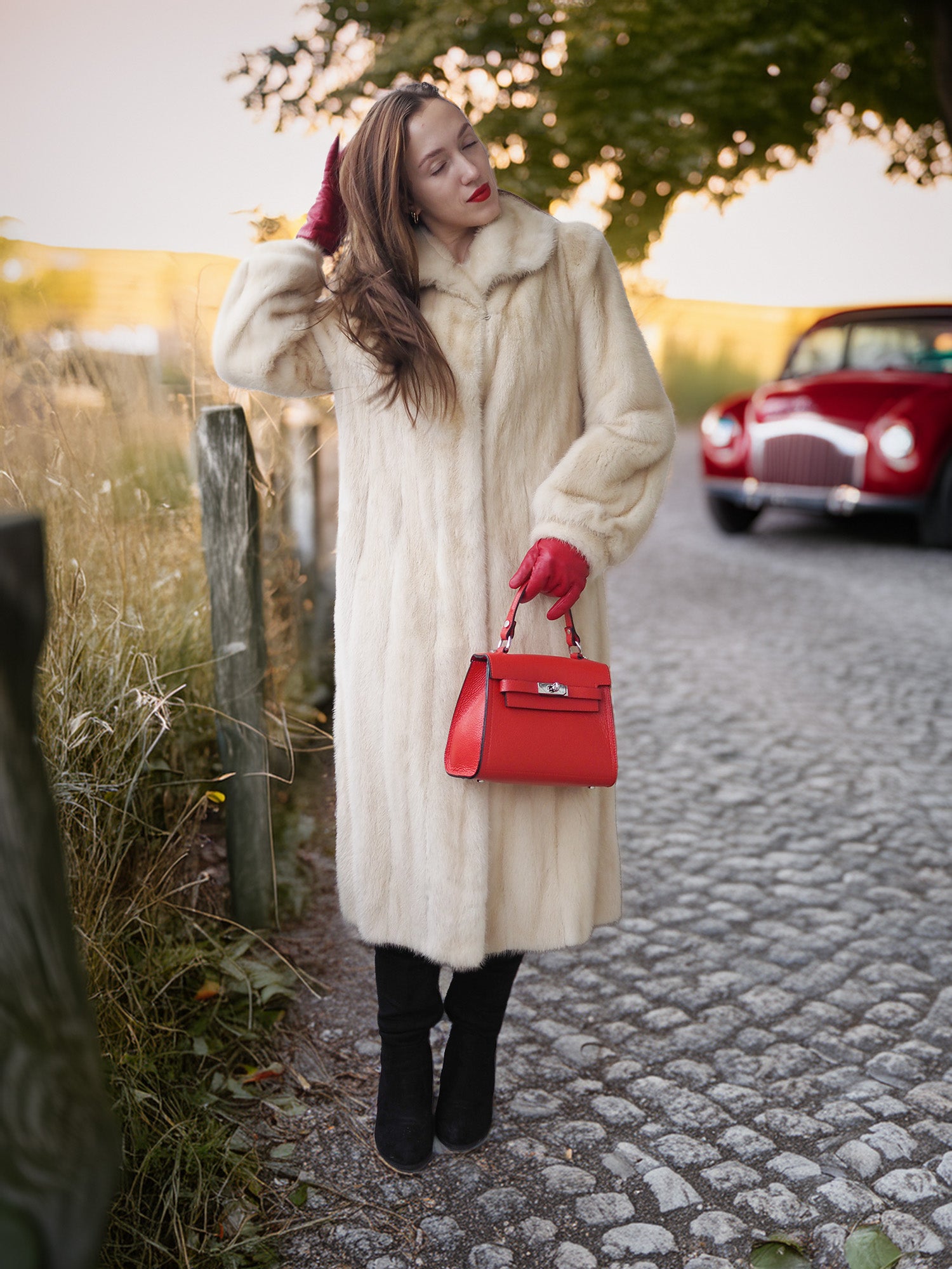 Woman in a long cream coat and red gloves holding a red handbag on a cobblestone street with a red car in the background.