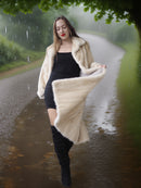 Woman in a long cream coat standing on a rainy path with greenery around.