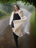 Woman in a long cream coat standing on a rainy path with greenery around.