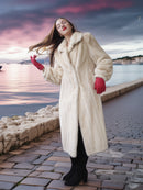 Woman in a fur coat and red gloves standing by a waterfront at sunset.