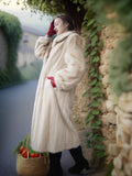 Woman in a long white fur coat holding a basket of vegetables, standing against a stone wall with greenery.
